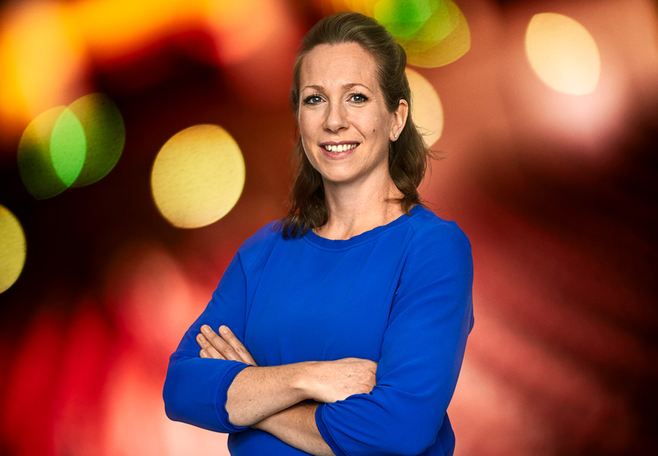 Kvinna med blå tröja och mörkt hår. Ler och står med armarna i kors. Fotografi.