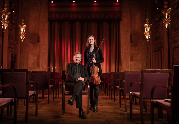 Man som sitter ned och kvinna som står bredvid med en viola. Fotografi.