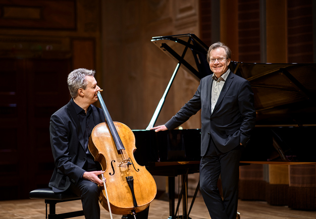 Two men, one with a cello and one standing against a piano. Photo