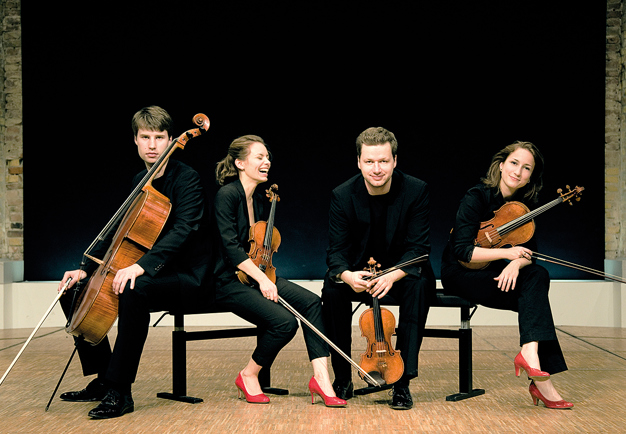 The quartet sitting on a bench indoors holding their instruments, one of them is laughing out loud with her eyes closed. Photography.