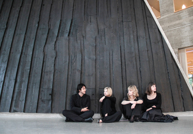 The quartet sitting talking to each other on the floor in front of a black wooden wall. Photography.