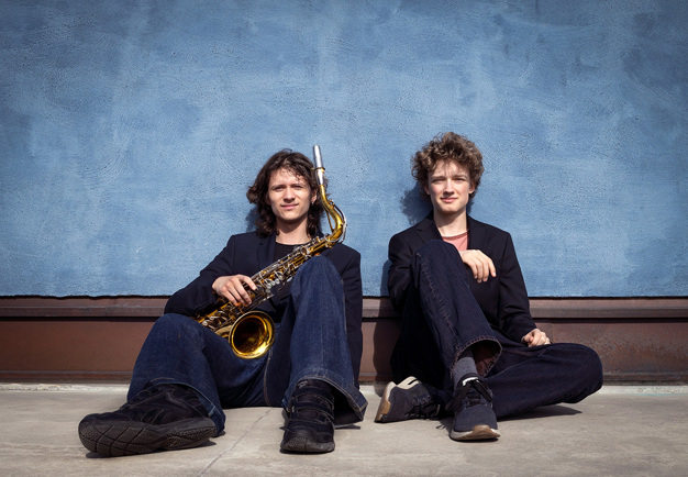 Två musiker som lutar sig mot en blå vägg. Fotografi.