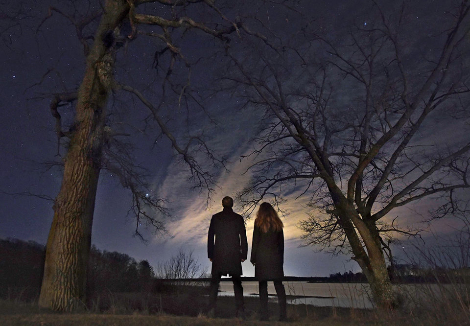 Två människor står med ryggarna mot kameran. De står utomhus under en stjärnhimmel. Stjärnorna poppar ut och blänker extra starkt. Fotografi. 