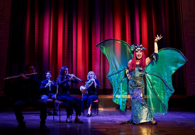 Drag queen in a green dragon dress, musicians in the background. Photo.