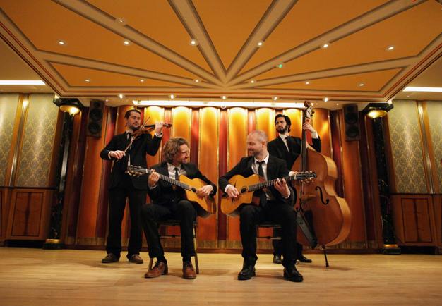 Gruppbild, alla med sina instrument i handen. En violinist, två gitarrister i förgrunden och en kontrabasist. Leende och varm känsla bland musikerna. Fotografi.