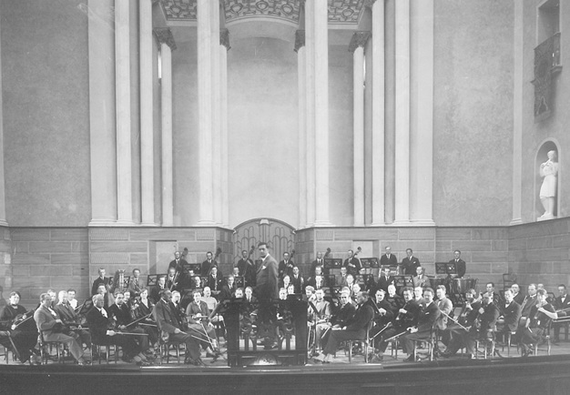 Gruppbild på orkestern i stora salen. Svart-vitt äldre fotografi.