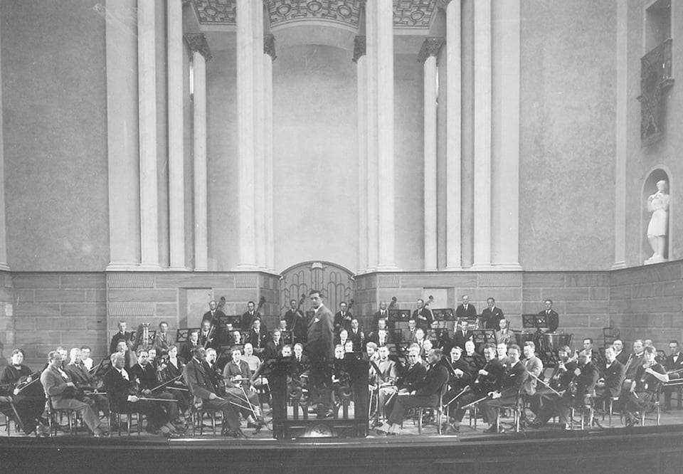Gruppbild på orkestern i stora salen. Svart-vitt äldre fotografi.