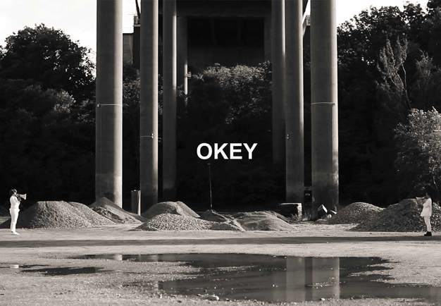 Black and white photo shot under a bridge, the word "okey" in white letters is placed in the middle of the photo. 