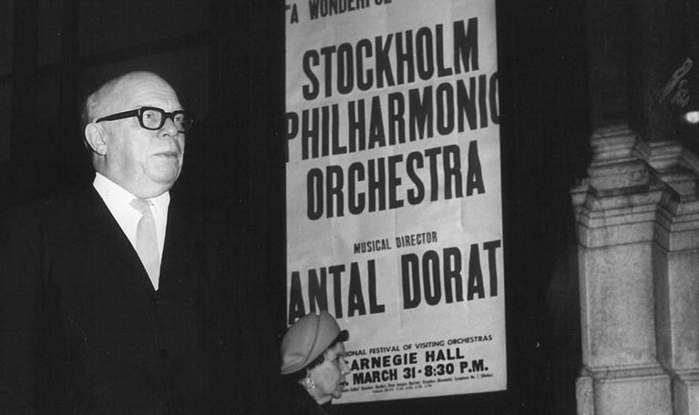 Black and white photograph of the director standing, posters of the orchestra's concert are clearly visible in the background.