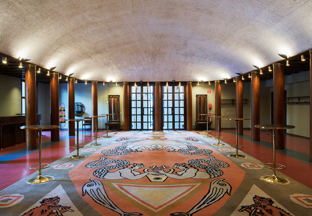 Et rum med välvt vitt tak, lampetter, träpelare och en stor figurativ matta på golvet. Fotografi.