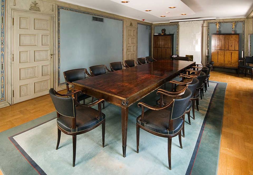 Ett pampigt mötesbord och 14 karmstolar skapat av Carl Milles mitt i bilden, ståtligt rum. I rummet står en kakelugn som har använts som kassaskåp förr i tiden. Fotografi.