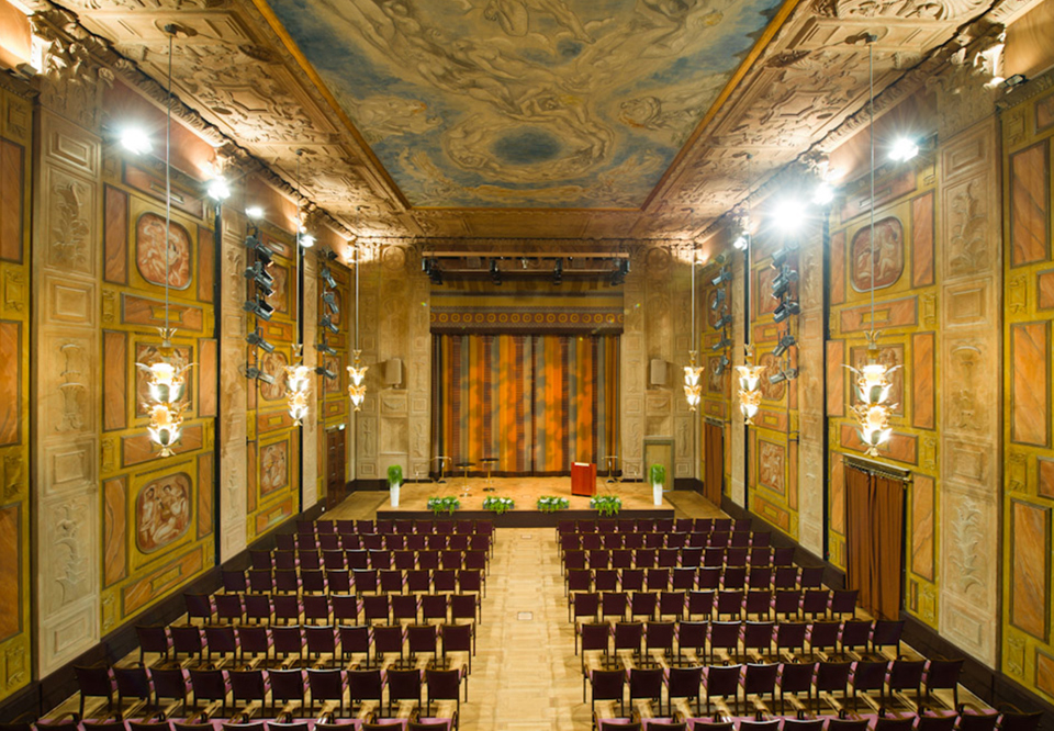 Grünewaldsalen sedd från längts bak i lokalen med talarstol på scen. Fotografi.