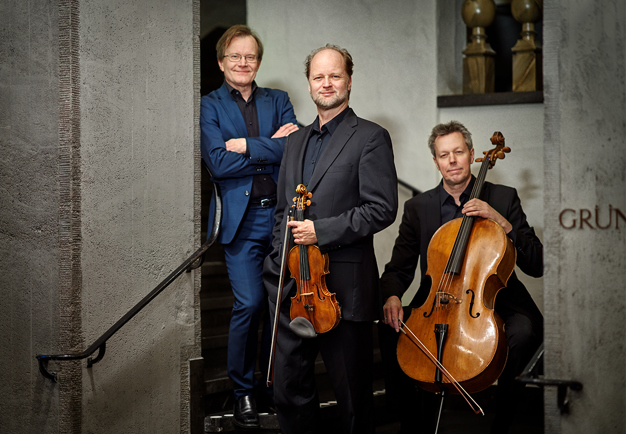 Three men, one of whom sits with his cello, one stands with his arms crossed and one stands and holds his violin. Photography.