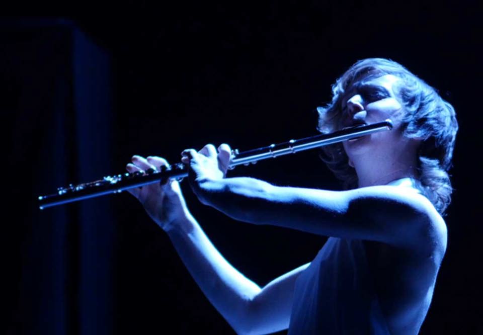 Claire Chase spelar på sin flöjt. Mörk bild med blått dramatiskt scenljus. Foto.