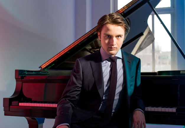 Sittande pianist med intensiv blick lutandes mot flygel. Daniil Trifonov. Fotografi.