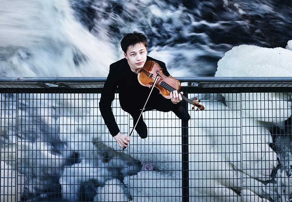 Kille som står vid ett vattenfall med sin violin. Fotografi.