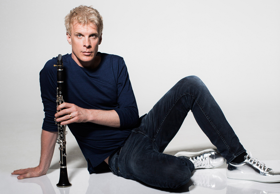Martin Fröst sitting on a white floor in front of a white background, holding the clarinet standing up on the floor in front of him.