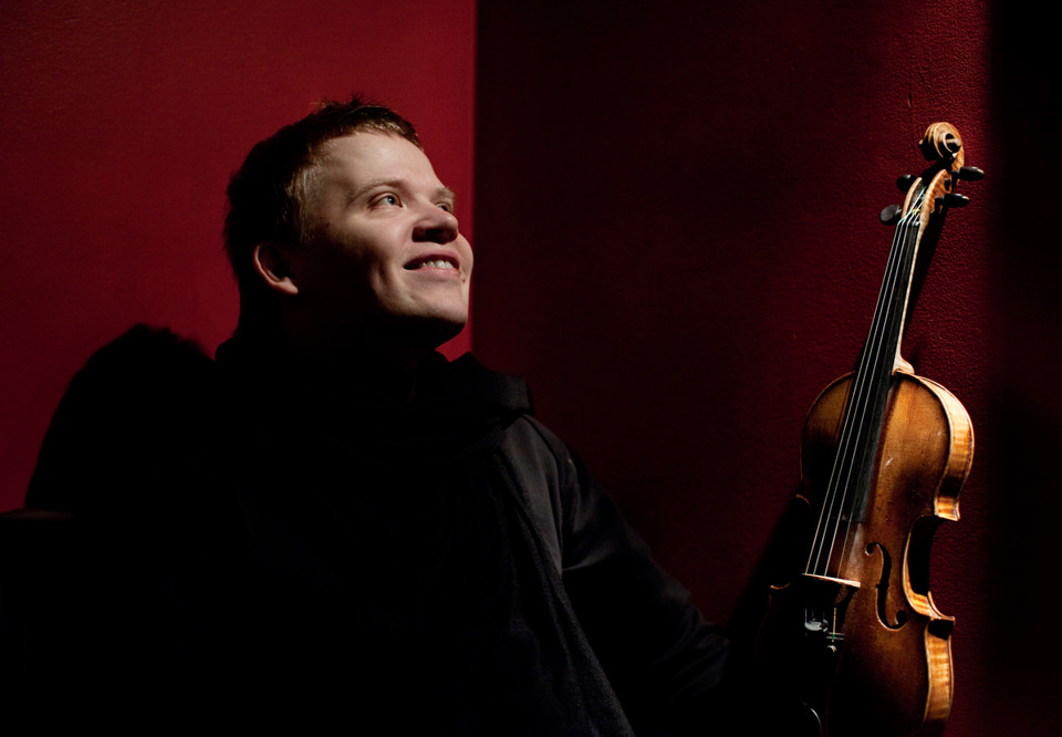 Glad violinist sittandes på stol mot röd bakgrund. Fiolen i knät. Pekka Kuusisto. Fotografi.