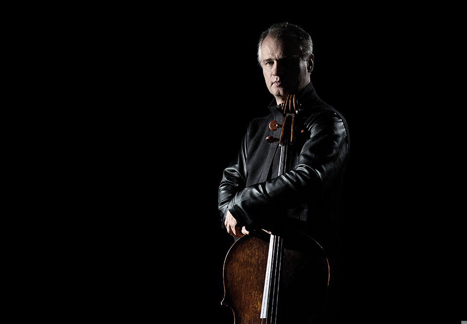 Cellist Torleif Thedéen standing up in sparse lighting, holding the cello beside him and looking at the camera with half his face in shadows. Photography.