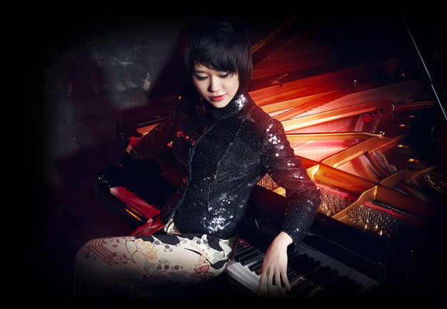 The pianist Yuja Wang sitting by an evocatively lit piano, with her back leaning onto the keyboard and her eyes looking down to the floor. Photography.