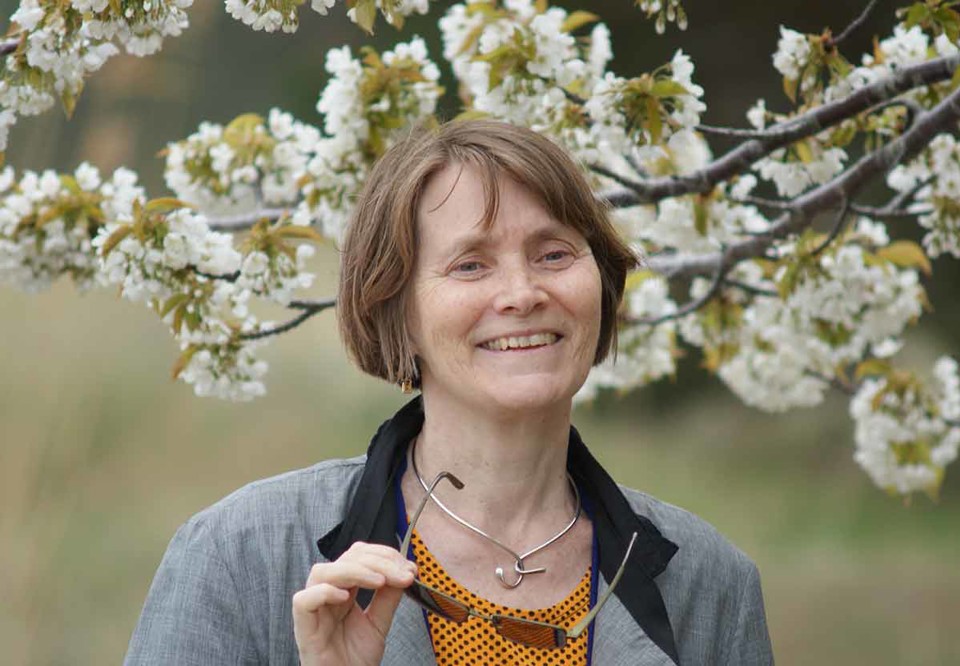 Cecilia håller i sina glasögon, står framför ett blommande träd i en blommig trädgår. Ser glad ut. Fotografi.
